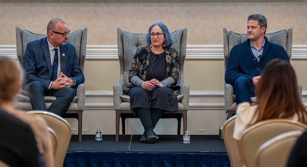 Three panelists, a man in a blue suit, a woman with blue hair, and a man in a blue cardigan, sit on a stage during a discussion.